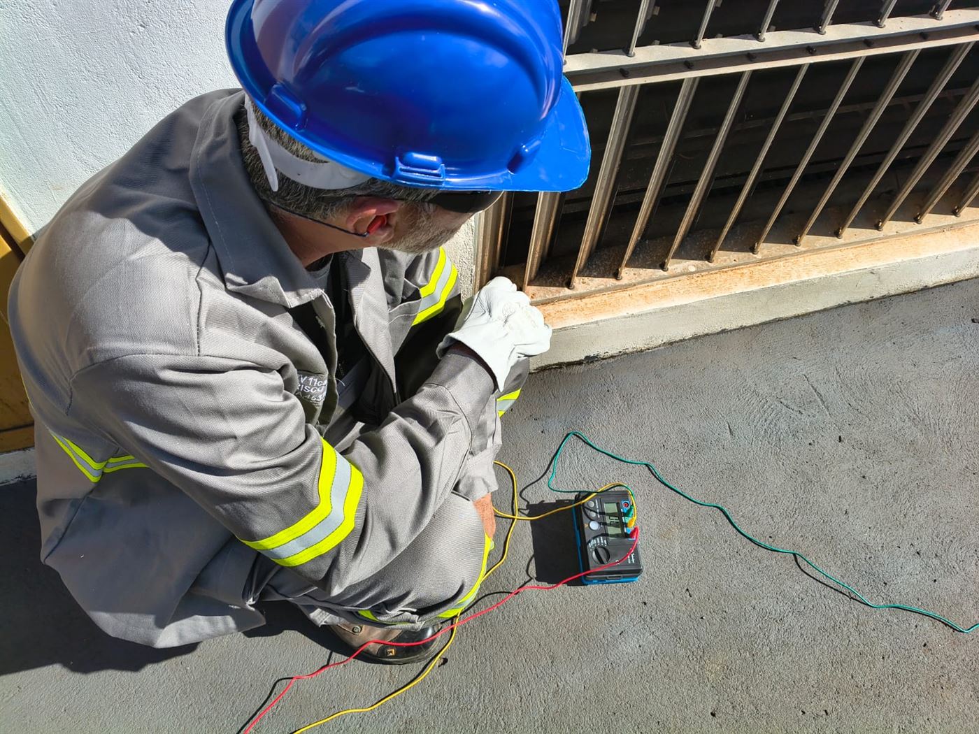 Técnico da IC agachado ao lado de terrômetro digital, com os cabos de ensaio ligados à caixa de inspeção