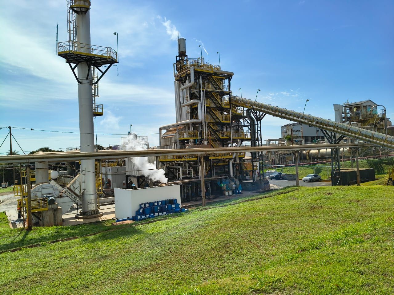 Planta agroindustrial com chaminés, torres e esteira transportadora, vista do gramado ao redor